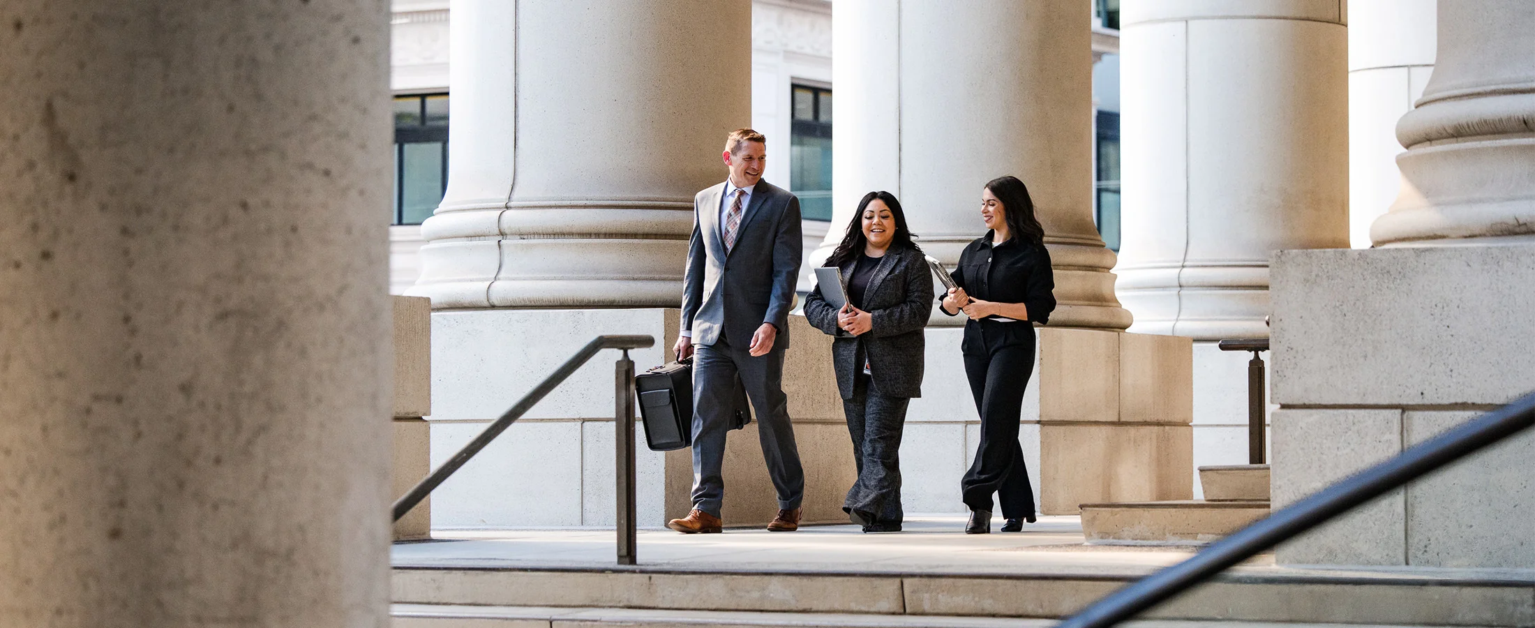 Three professionals walk together through a spacious, modern lobby or corridor featuring tall stone columns and large windows. Each person carries a rolling suitcase or briefcase, suggesting business travel or arrival for a meeting. The setting is bright.