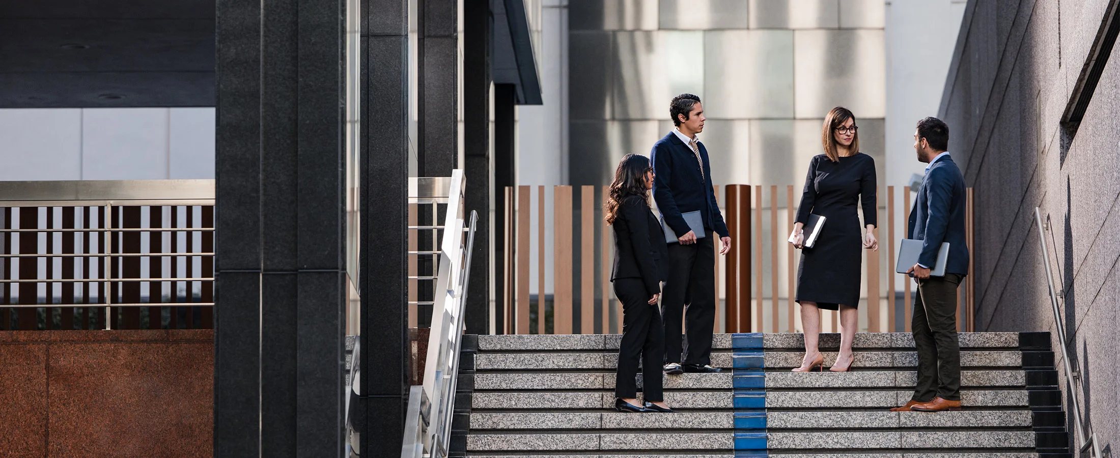Four professionally dressed people stand on wide stone steps outside a modern office building, holding laptops and folders while engaged in conversation. The scene features contemporary architecture with glass, metal, and dark stone surfaces, and natural