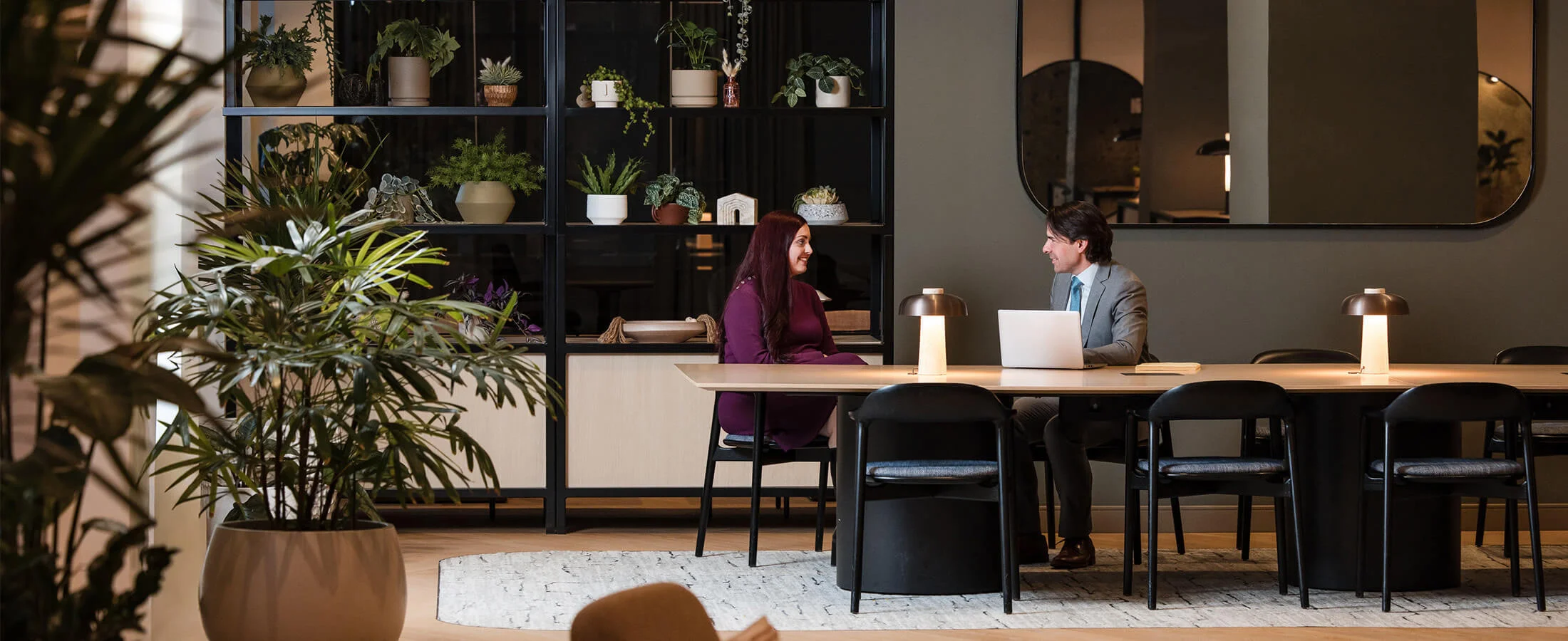 A modern café-style workspace with a long wooden counter and high stools sits in a warmly lit interior. Two people are positioned behind the counter working with laptops, while the foreground includes green plants and a light-colored sofa with cushions.