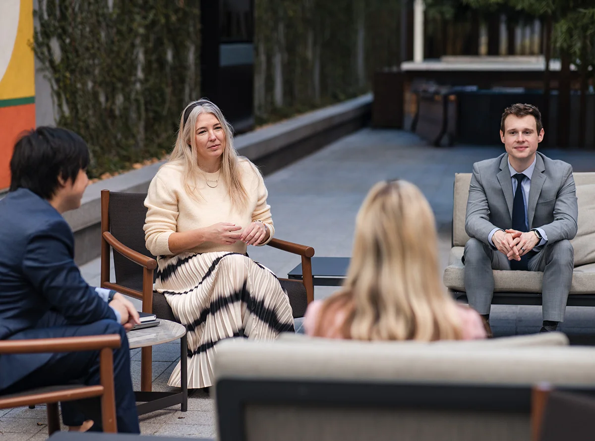 Four professionals sit and converse at a casual, comfortable, professional environment, sitting in short chairs and natural lighting.