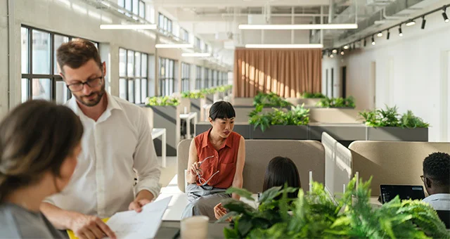A bright, modern open office with long rows of desks, overhead lighting, and large windows along one wall. Several individuals collaborate in small groups; in the foreground, one person reviews papers while another sits and gestures during a conversation.