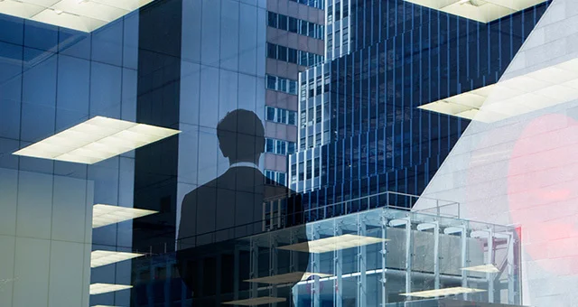 Reflection of a modern office interior with ceiling lights overlaid on glass high‑rise buildings, featuring the silhouette of a person standing and looking outward, creating a layered urban and corporate scene.