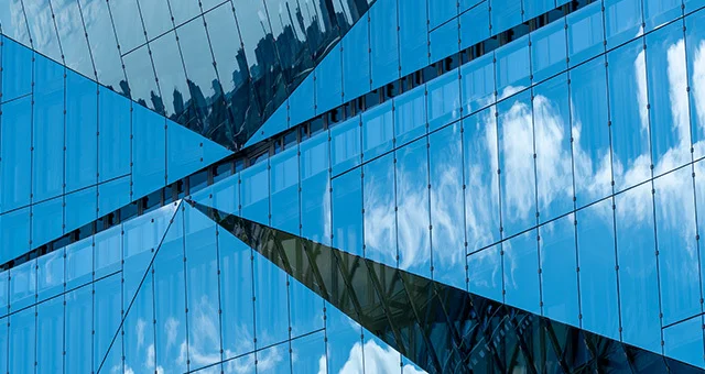 Close-up of a modern glass building exterior with geometric lines and reflective panels, mirroring blue sky and clouds with intersecting architectural shapes.