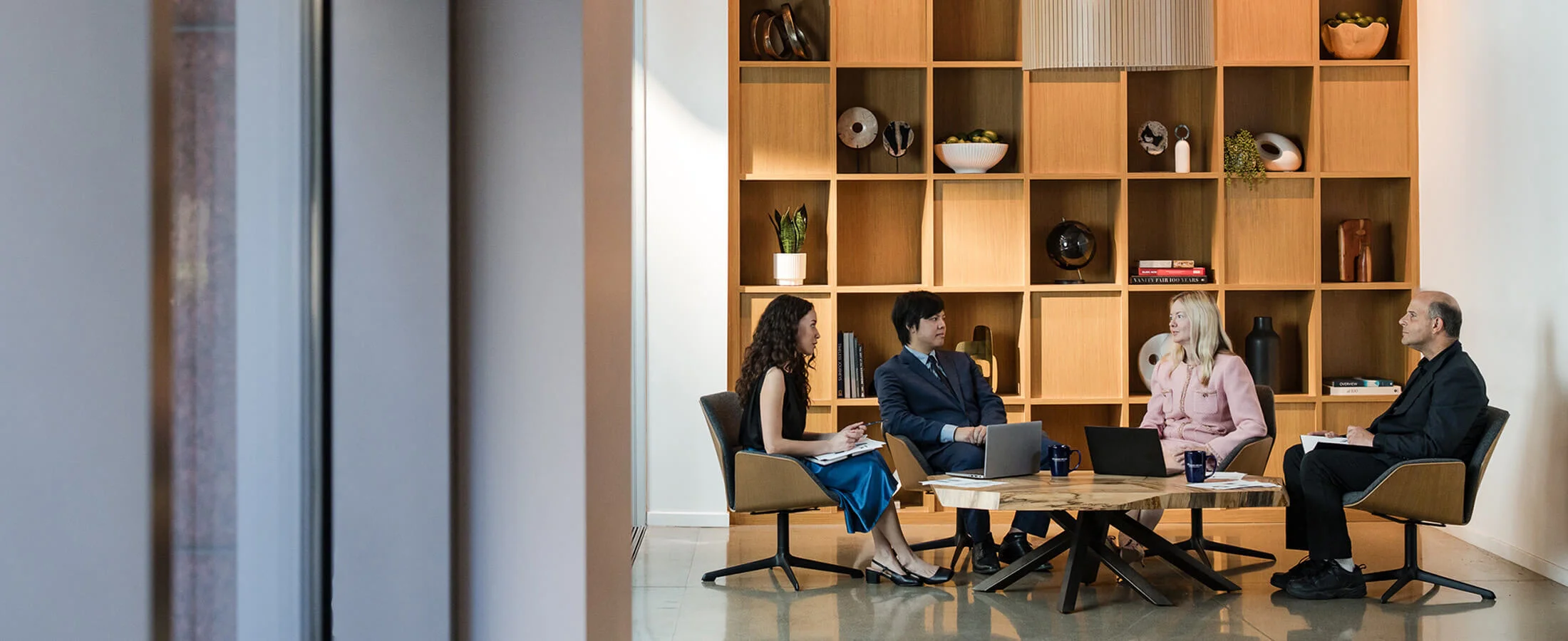Four people sit around a round wooden table in a modern office lounge, engaged in a discussion with open laptops and notebooks. Behind the group is a large floor-to-ceiling wooden shelving wall displaying books, plants, pottery, and decorative objects. Na