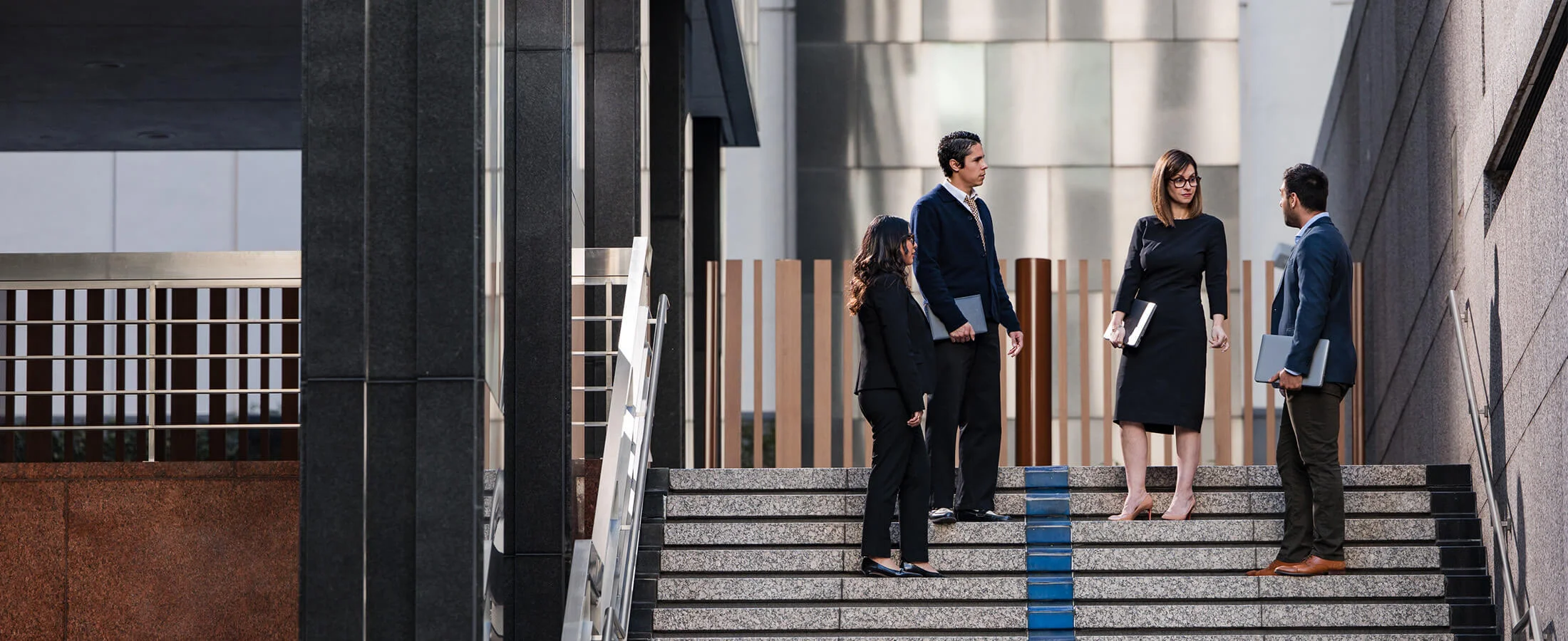 Four professionally dressed people stand on wide stone steps outside a modern office building, holding laptops and folders while engaged in conversation. The scene features contemporary architecture with glass, metal, and dark stone surfaces, and natural