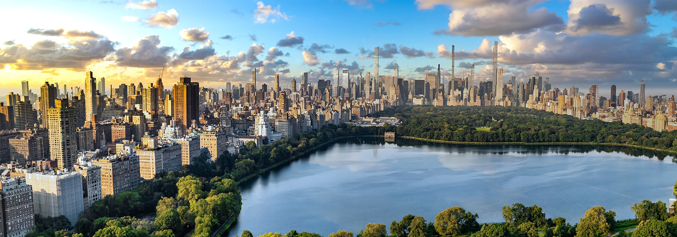 “Panoramic view of Manhattan skyline at sunset, with Central Park’s tree-lined reservoir in the foreground and tall skyscrapers rising behind under a partly cloudy sky.