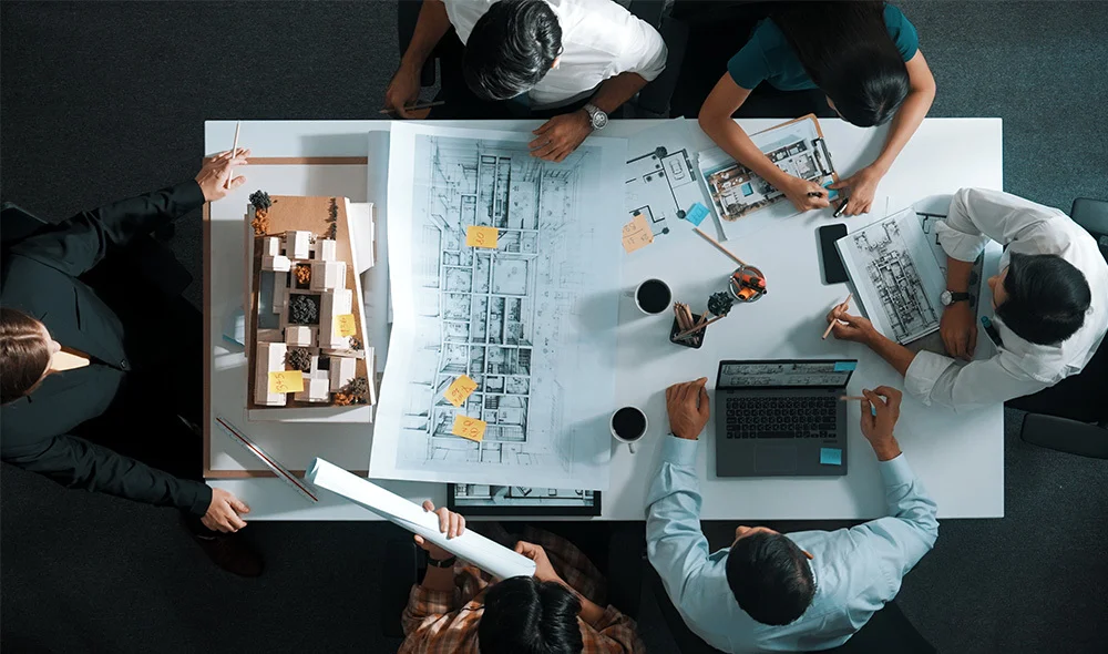 Overhead view of a formal business meeting around a rectangular wooden table. Several individuals in business attire are seated around the table, each with documents, notebooks, or devices placed in front of them. One person is at the head of the table.