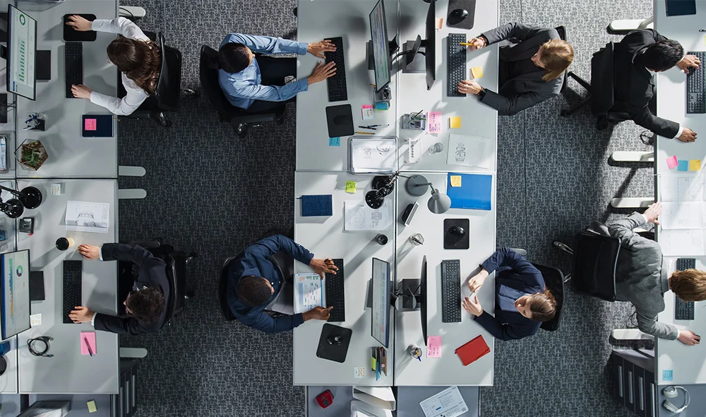 An overhead view of a busy, modern office environment shows multiple desks arranged in a grid-like layout. Each workstation is equipped with computer monitors, keyboards, office supplies, and paperwork. Several individuals are seated or moving.