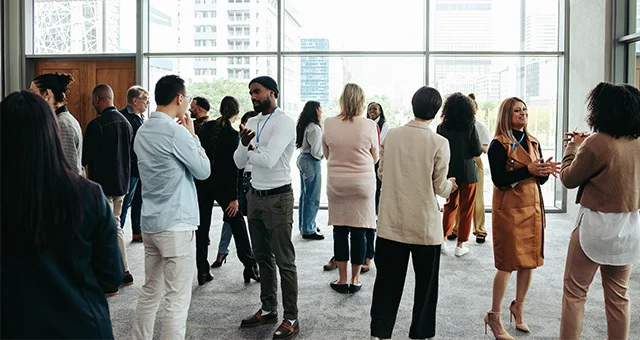 Group of adults wearing name badges gathered in a bright, modern indoor space, standing and talking in small groups near large windows overlooking a city skyline.