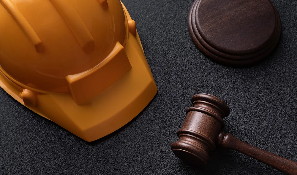 Close-up of a yellow construction hard hat placed next to a wooden gavel and round sound block on a dark textured surface, suggesting a connection between workplace safety regulations and legal proceedings.