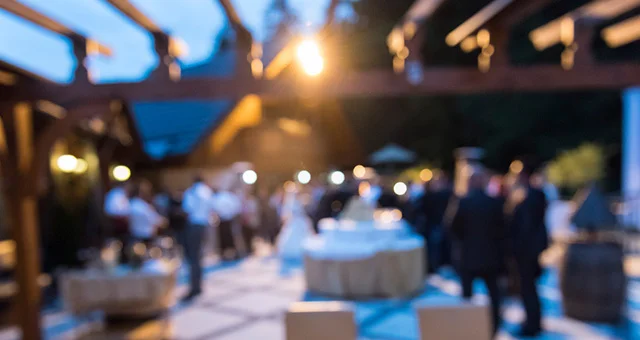Softly blurred evening scene of an outdoor event with guests gathered around cocktail tables under warm lights, suggesting a reception or social gathering in a courtyard setting.