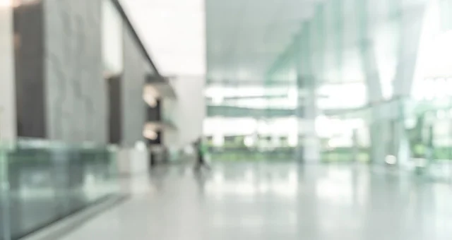 A bright, modern office lobby or corridor features a spacious, open layout with glossy floors and large glass windows allowing natural light to flood the space. In the distance, several people walk through the corridor, appearing small.