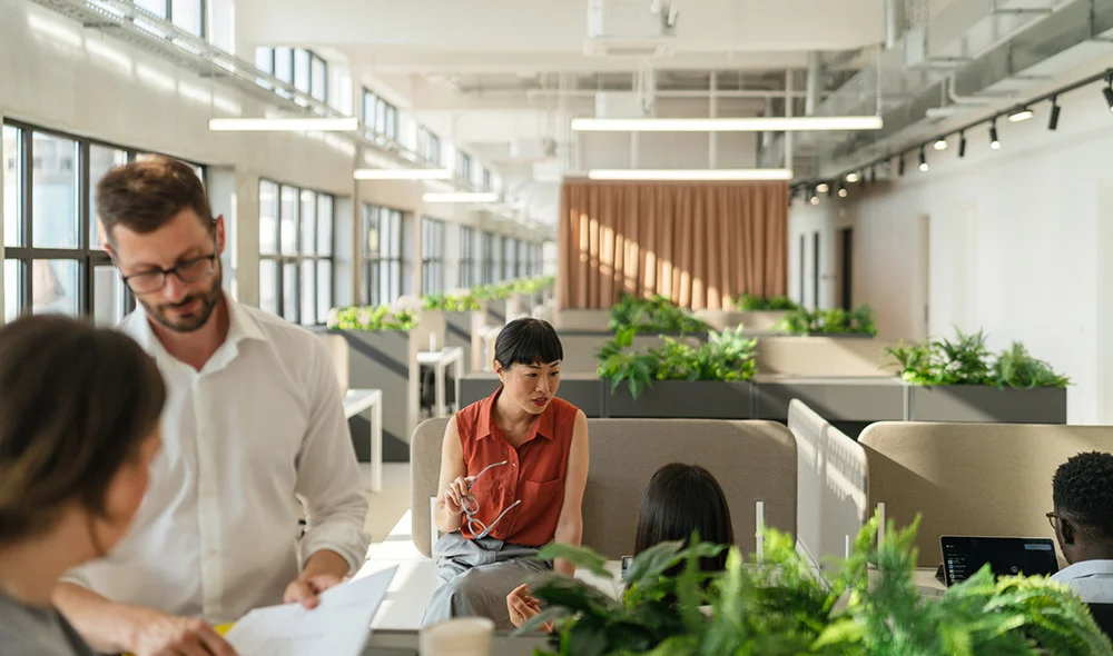 Open, modern office workspace with large windows and rows of plants, where several people collaborate around a seating area while reviewing documents and discussing work.