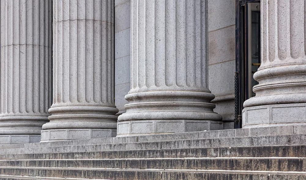 Stone steps leading up to a neoclassical building with large, fluted columns and a partially visible doorway, featuring textured gray stone and architectural detail suggestive of a courthouse or civic building.