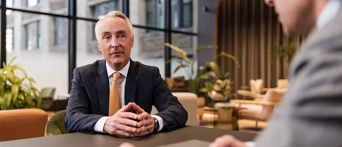 A professional office or meeting space with a person in the foreground seated at a table, wearing a dark suit and a light-colored shirt. The person’s hands are clasped together on the table, suggesting engagement in a conversation or meeting.