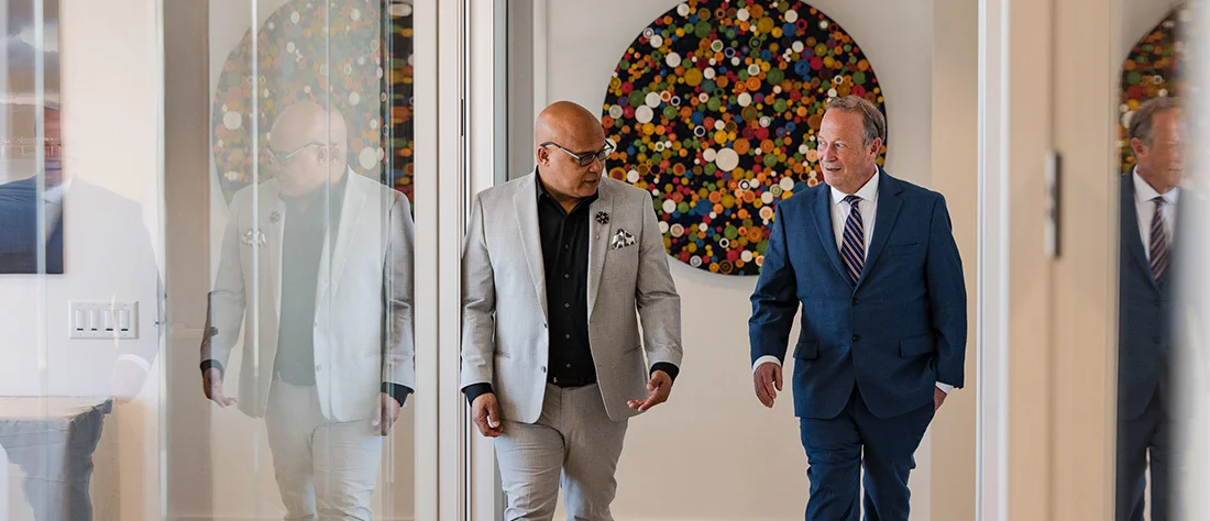 Two adults in business attire walk side by side through a bright, modern interior hallway. One adult on the left wears a light gray suit with a white top and carries a bag, while the adult on the right wears a dark blue suit with a white shirt.
