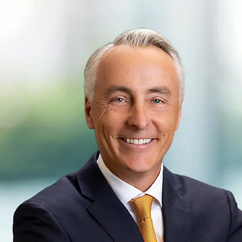 Professional headshot of Kevin O'Hagan in a dark suit and white shirt with an orange tie, facing forward with a neutral posture against a softly blurred office background with large windows and cool-toned light.