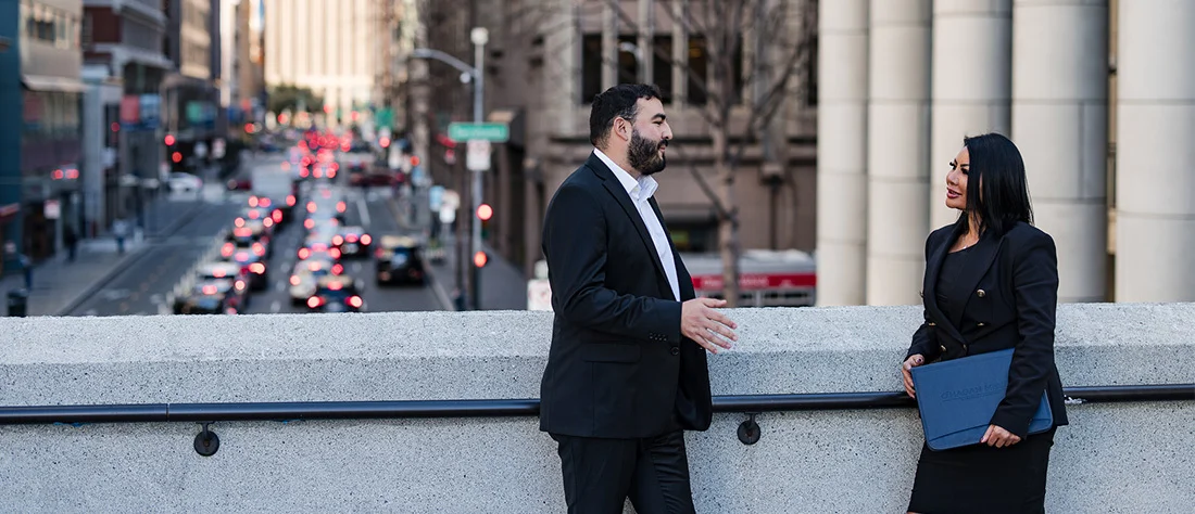 Two professionals stand on a balcony overlooking a city street filled with cars and traffic below. One individual leans against a railing while looking out over the street, wearing a dark suit, while another individual in business attire is also in view.