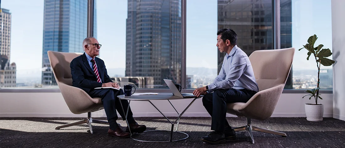 Wide shot of a modern office lounge area with two professionals seated across from each other on light-colored couches. Between them is a small table, creating a conversational setup.