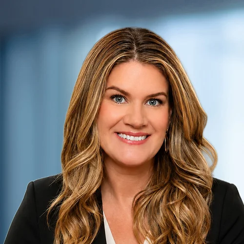 Professional headshot of Jamie Filipovic with long, wavy light brown hair, wearing a black blazer over a light-colored top. The person is facing forward and looking toward the camera. The background is softly blurred in cool blue and gray tones, suggestin