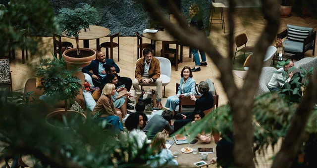 A group of people sits in a circle of chairs in a spacious indoor atrium with wooden flooring and abundant greenery. Small round tables, potted trees, and additional seating surround the gathering, while large plants and a tree trunk partially frame the s