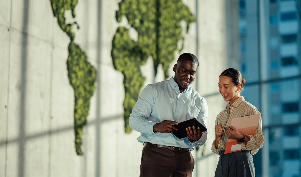 Two professionals stand together in a bright, modern office environment near large glass windows. One individual holds an open laptop angled toward the other, who looks at the screen while holding papers or a notebook.