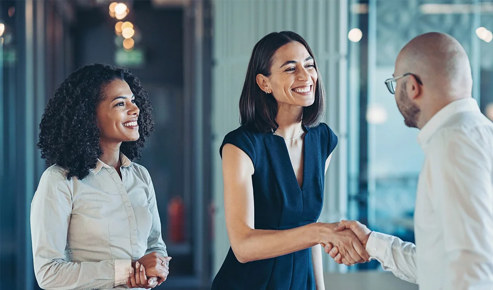Two professionals stand facing a third person in a modern office environment, engaged in conversation. One person in the center holds a tablet or folder, while the other person beside them stands with arms relaxed at their sides.