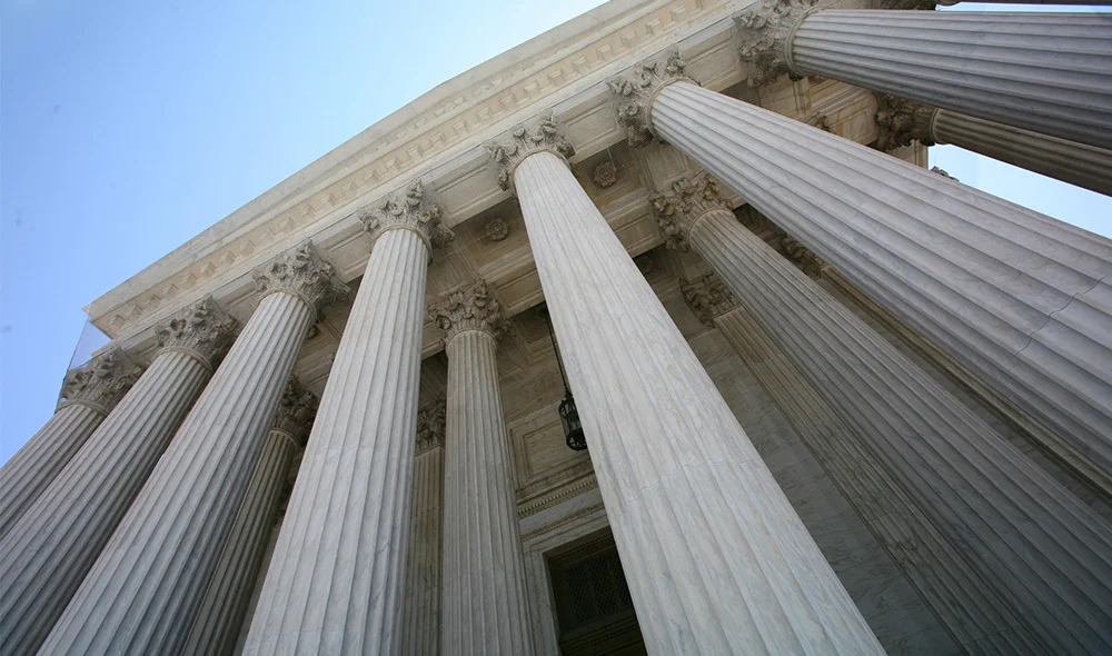 A low-angle view of tall, white neoclassical columns rising vertically against a clear blue sky. The columns feature smooth shafts with simple capitals and are arranged in a symmetrical row, creating a strong sense of height, structure, and arch.