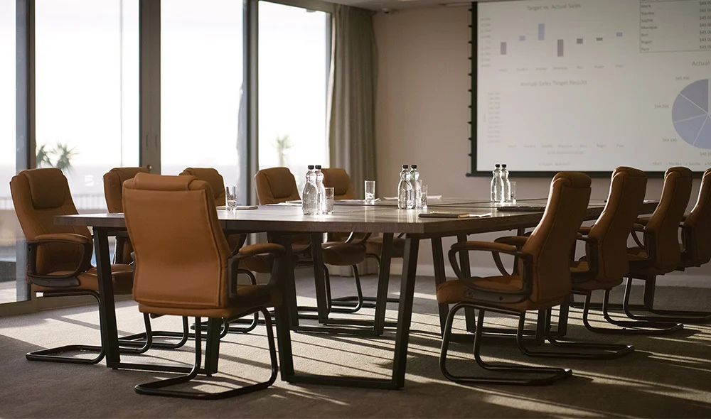 Conference room with a long wooden table surrounded by brown leather swivel chairs, set with water bottles and glasses. Floor-to-ceiling windows let in natural light, and a projection screen at the front displays charts and graphs for a presentation.