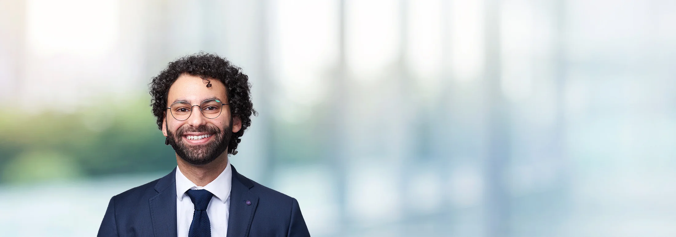 A smiling man with curly dark hair and a beard, wearing glasses, a navy suit, white shirt, and tie, posed against a softly blurred office background.