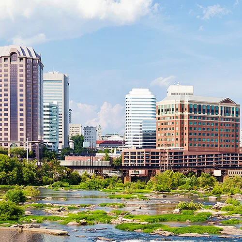Wide view of downtown Richmond, Virginia, with mid- and high-rise office buildings rising behind a river filled with rocks and greenery, and a concrete bridge crossing from the left under a bright, partly cloudy sky.