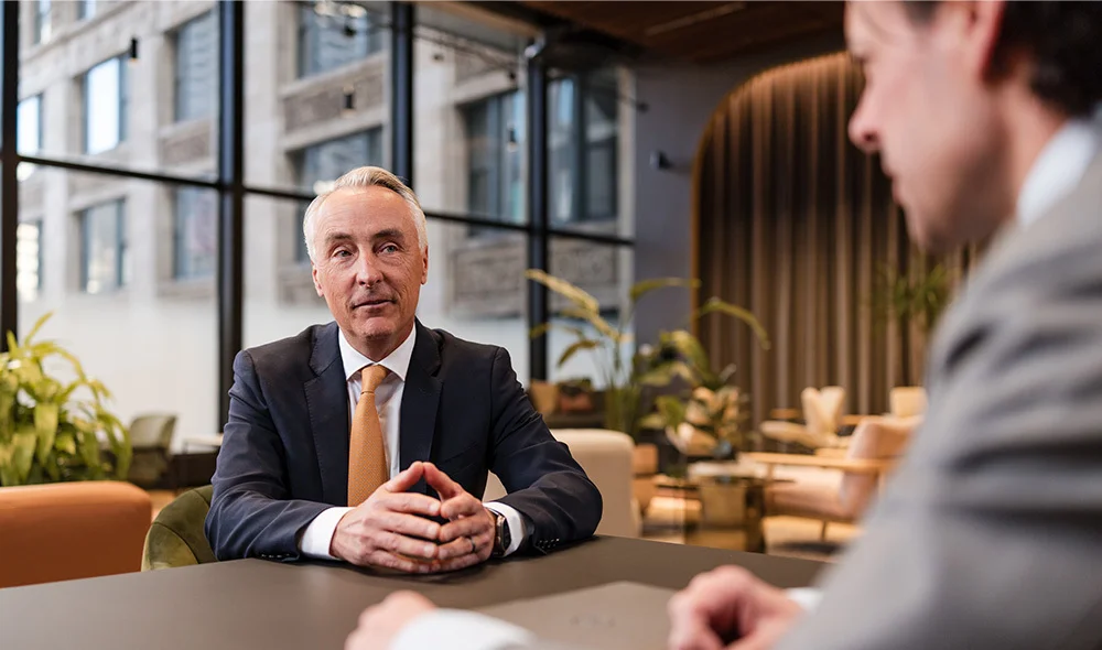 Two individuals sit across from each other at a wooden table in a modern office, engaged in a professional discussion. One individual in the foreground is partially visible, while the other individual faces forward and leans slightly toward the table.