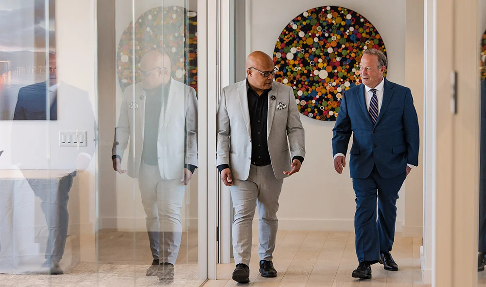 Three professionally dressed individuals walk together in a bright indoor corridor with large windows and light-colored walls. One individual walks slightly ahead while the other two walk side by side, all carrying themselves in a businesslike manner.