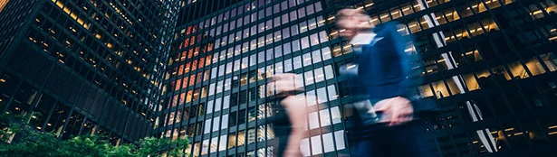 Blurred professionals walk through a downtown business district surrounded by tall office buildings, conveying the fast-paced nature of legal and corporate work.