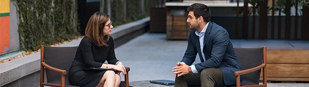 Two professionals seated across from each other in an outdoor office courtyard, leaning forward and engaged in conversation with a tablet placed on a low table between them.