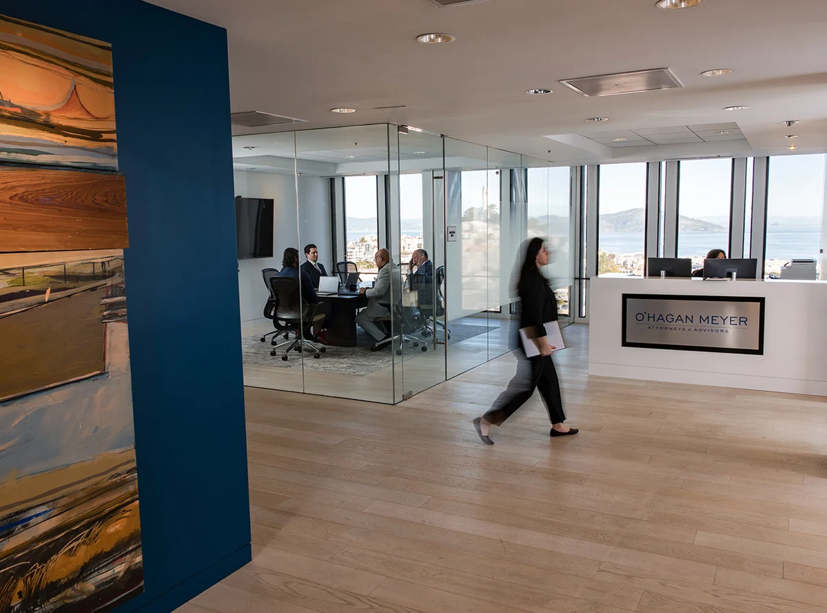 Modern corporate office reception area with light wood floors and floor‑to‑ceiling windows overlooking a waterfront city view. A white front desk displays the text “O’Hagan Meyer Attorneys + Advisors.” A glass conference room sits to the left with several