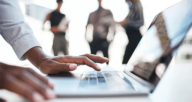Close-up of a person’s hand using a laptop keyboard in a bright office setting, with several people standing and conversing in the blurred background, suggesting a collaborative work environment.