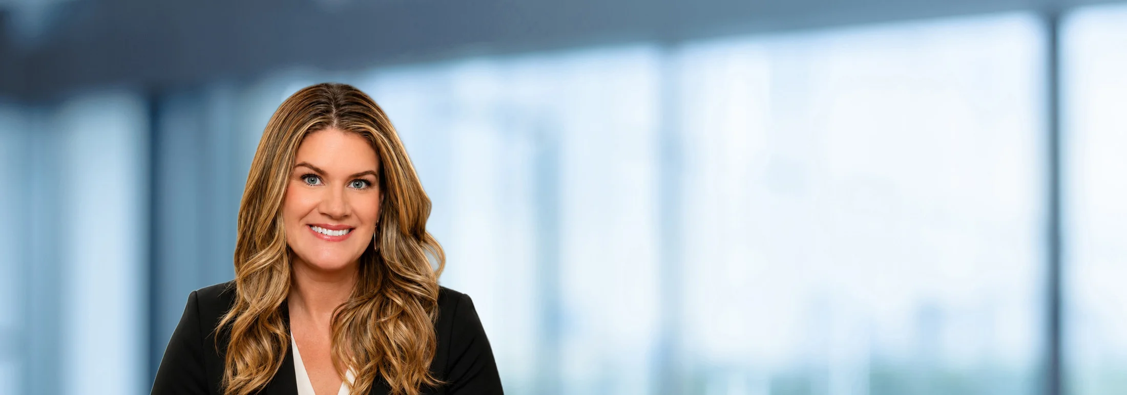 Professional headshot of Jamie Filipovic with long, wavy light brown hair, wearing a black blazer over a light-colored top. The person is facing forward and looking toward the camera. The background is softly blurred in cool blue and gray tones, suggestin