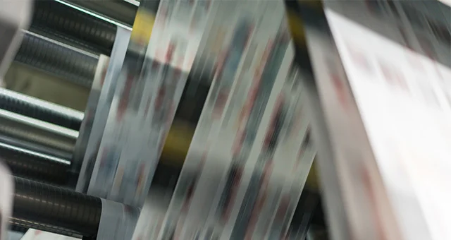 Close-up of a printing press in motion, with sheets of printed material moving rapidly through metal rollers, creating a blurred effect that conveys speed and industrial production.