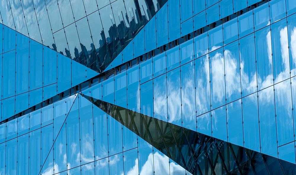 Close-up of a modern glass building exterior with geometric lines and reflective panels, mirroring blue sky and clouds with intersecting architectural shapes.