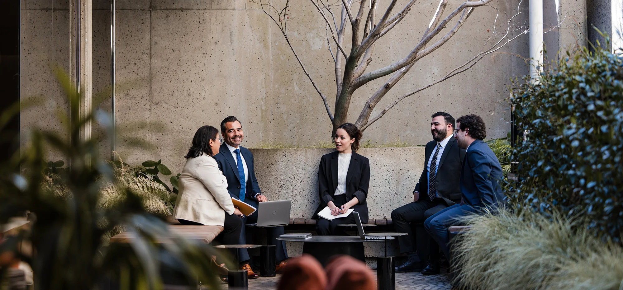 A small group of professionals stands outdoors near a building with bare trees and landscaping visible around them. One person holds a tablet or clipboard and directs attention toward it while others gather closely, looking at the device.