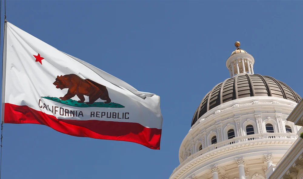 View of a domed government building with classical architecture, featuring a large white dome rising above a circular colonnade. A flagpole displays the California state flag, which includes a red stripe, a bear emblem, and the word “California".