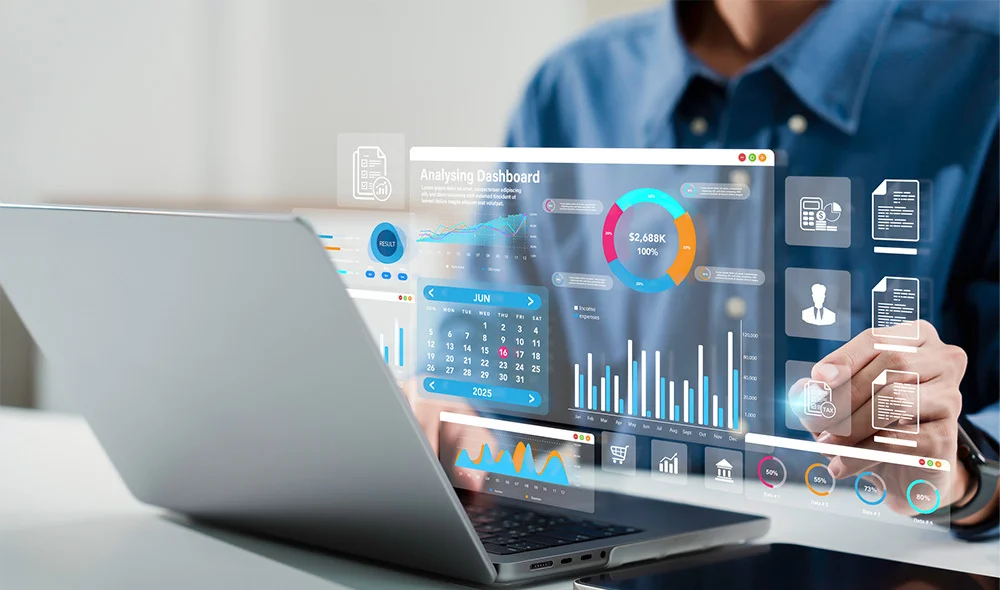 Close-up of a person in business attire working on a laptop at a desk, with a transparent digital overlay showing colorful data charts and graphs. The screen displays bar charts, line graphs, and numeric indicators in bright colors.